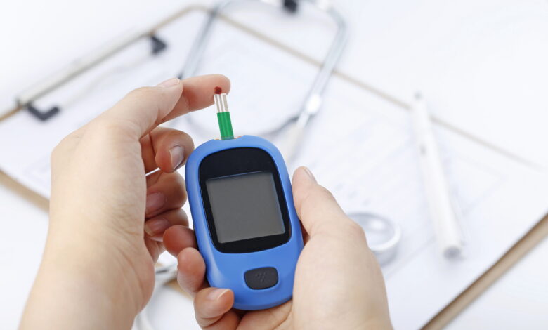 Hand holding a blood glucose meter measuring blood sugar, the background is a stethoscope and chart file